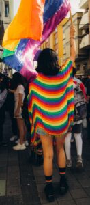Von hinten ist eine Frau mit schwarzem, langem Haar zu sehen. Sie trägt Shorts und einen Regenbogenponcho. Sie wedelt mit einer großen Regenbogenflagge.