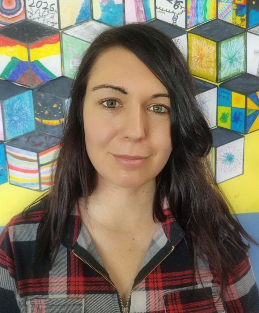 Anne lächelt in die Kamera. Sie hat langes, schwarzes Haar und grüne Augen. Sie trägt ein kraiertes Shirt.