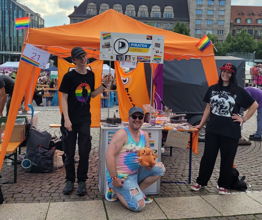 Tim von den Piraten Sachsen sowie Anne und Marcel vor unserem orangefarbenen und bunt dekorierten Infostand auf dem CSD Leipzig. Alle drei lächeln direkt in die Kamera.